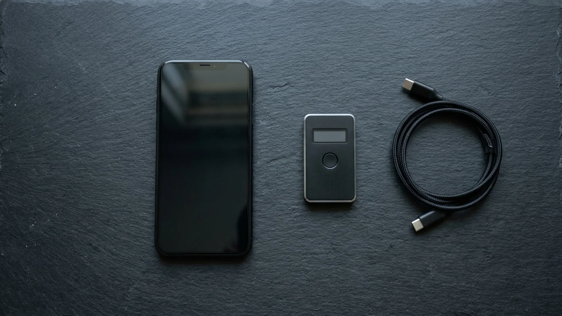 A phone and a hardware wallet side by side on a dark surface, cables coiled nearby, showing two very different devices for holding keys