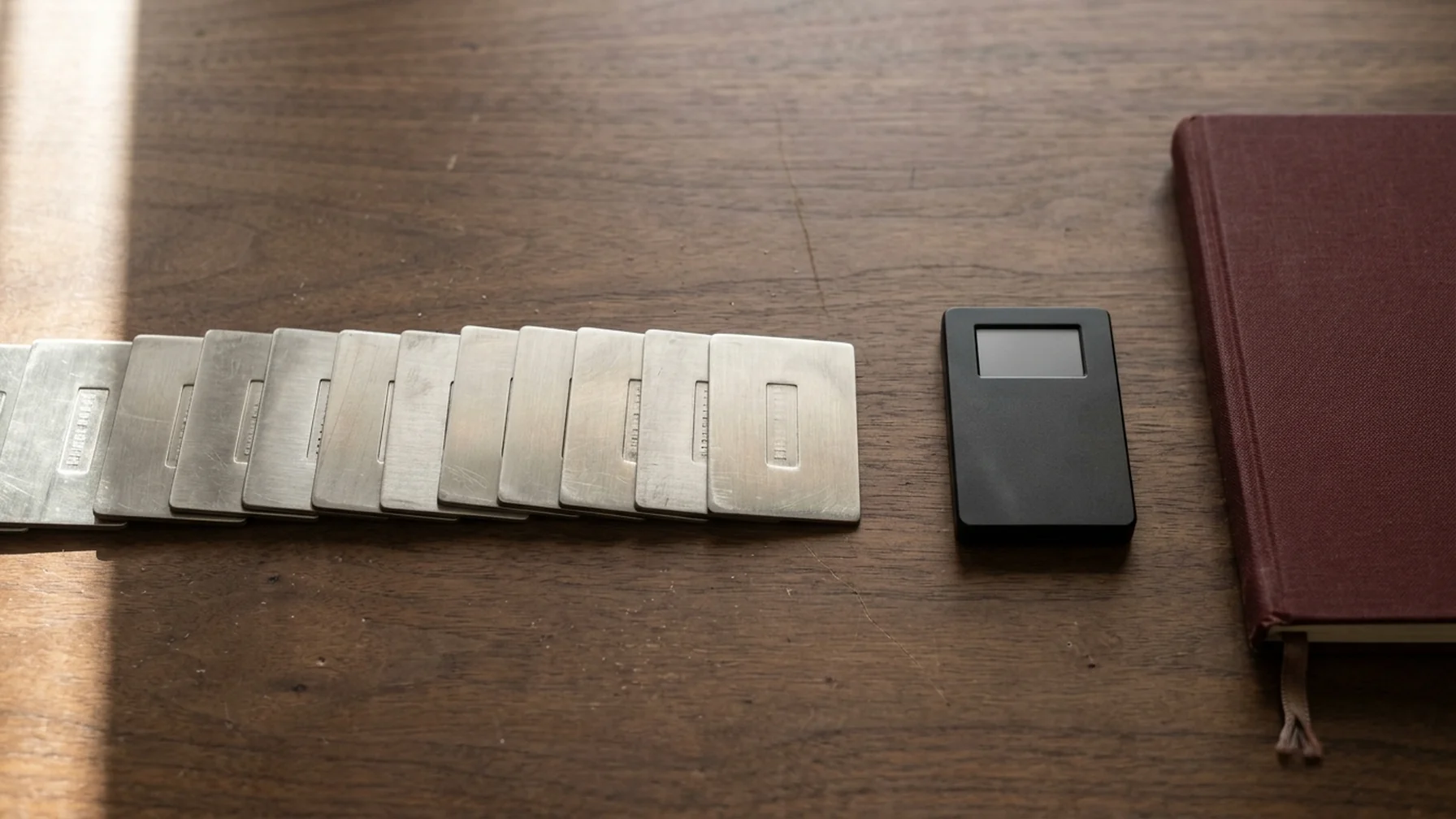 A seed phrase stamped onto metal plates laid on a wooden desk beside a hardware wallet and a notebook, lit by window light